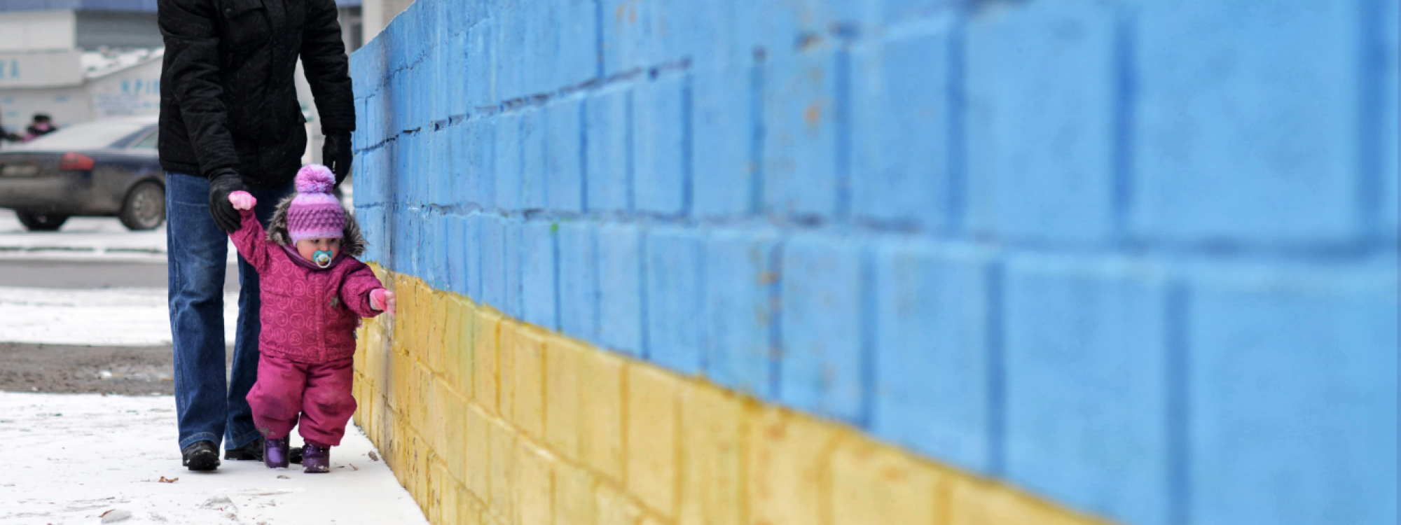 Winter scene: Man walking with small toddler by a brick wall painted in Ukrainian colours